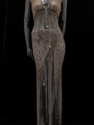 Elegant metallic gown with intricate beadwork and dangling chain details on a black background.