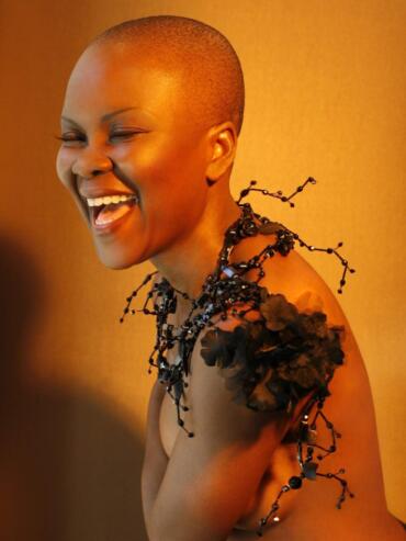 A joyful woman with a buzzed hairstyle wearing a unique, avant-garde black jewelry piece and floral-details on her shoulder.