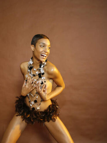 A woman posing confidently in a feathered skirt and layered jewelry against a brown background.