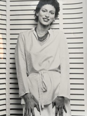 Black and white portrait of a woman with short, curly hair, smiling, wearing a light-colored, belted dress and layered necklaces, standing in front of a wooden slat background.