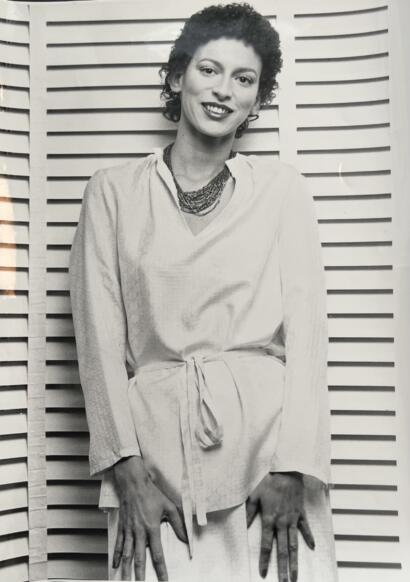 Black and white portrait of a woman with short, curly hair, smiling, wearing a light-colored, belted dress and layered necklaces, standing in front of a wooden slat background.