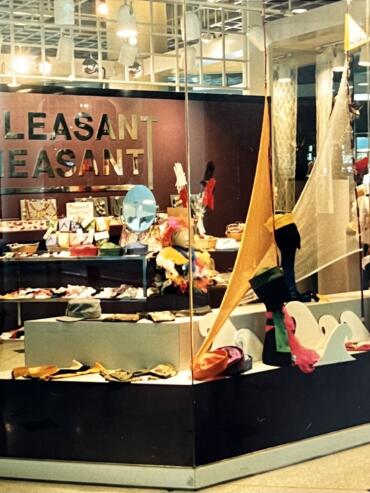 Display window of a store featuring colorful fashion accessories and artful textiles, with "PLEASANT" sign in the background.