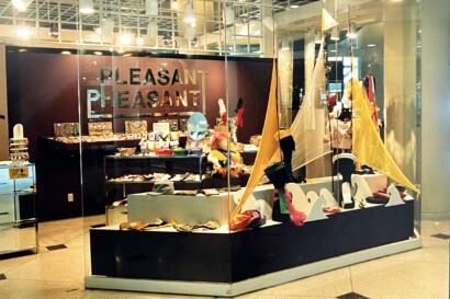 Display window of a store featuring colorful fashion accessories and artful textiles, with "PLEASANT" sign in the background.