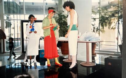 Three women in eclectic, colorful outfits converse indoors with large windows and potted plants behind them.