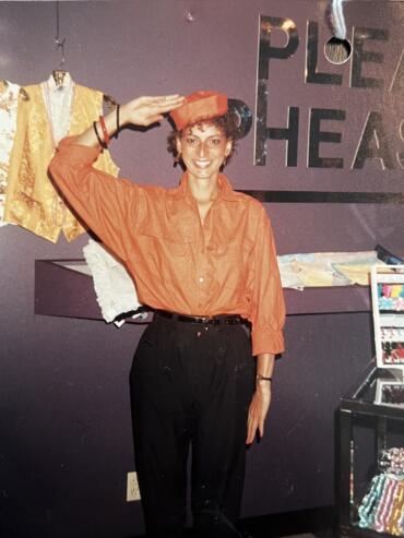 Woman in orange shirt saluting at a craft fair, with "PLE THE" visible in the background.