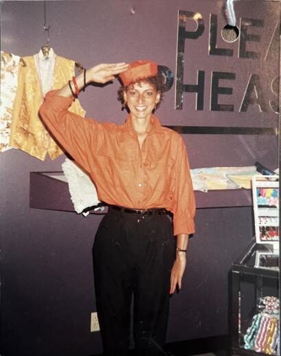 Woman in orange shirt saluting at a craft fair, with "PLE THE" visible in the background.