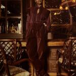 Elegant woman in a deep wine-toned, tiered dress with a tied waist, posing in a warmly lit, vintage-style interior.