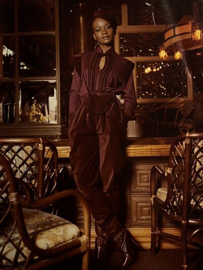Elegant woman in a deep wine-toned, tiered dress with a tied waist, posing in a warmly lit, vintage-style interior.
