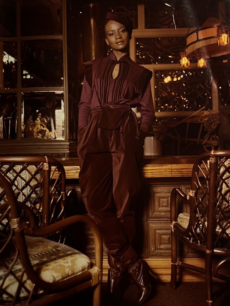 Elegant woman in a deep wine-toned, tiered dress with a tied waist, posing in a warmly lit, vintage-style interior.