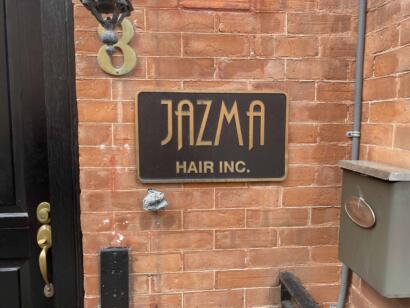 Sign for JAZMA Hair Inc. mounted on a brick wall outside a building.