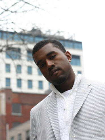 Portrait of a young man in a striped blazer and textured white shirt outdoors against a cityscape.