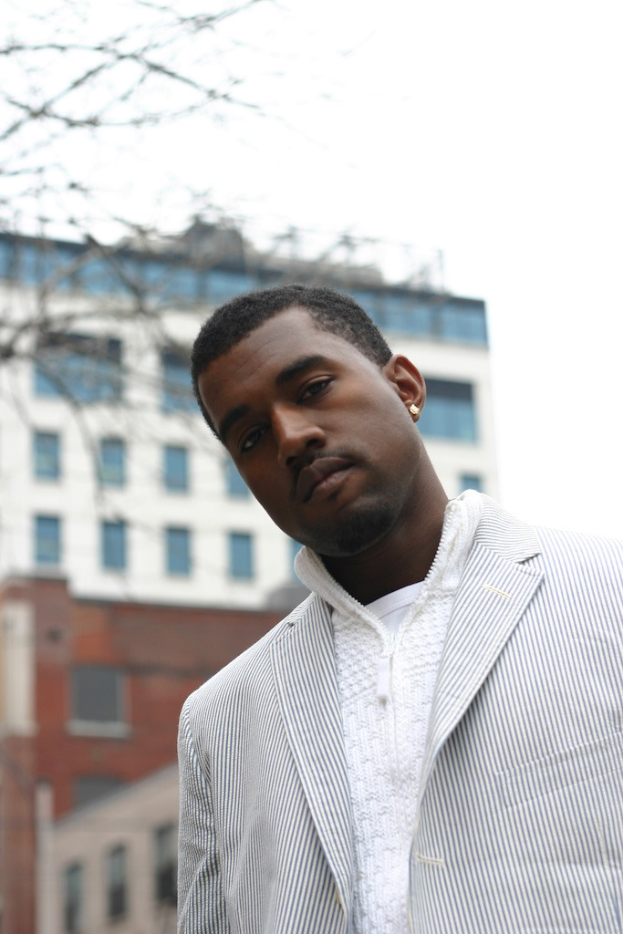 Portrait of a young man in a striped blazer and textured white shirt outdoors against a cityscape.