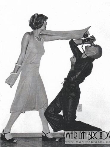 A woman in a dress and heels pushes a man with a camera in a playful pose.