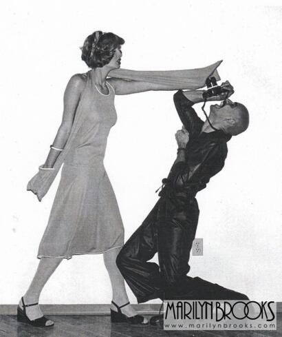 A woman in a dress and heels pushes a man with a camera in a playful pose.