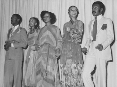 Black and white photo of five diverse individuals in elegant clothing standing on stage.