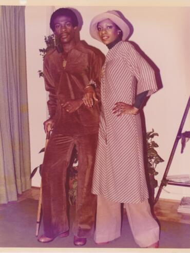 Two women pose indoors, one in a brown velvety suit and the other in a striped dress with a wide-brim hat.
