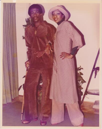 Two women pose indoors, one in a brown velvety suit and the other in a striped dress with a wide-brim hat.