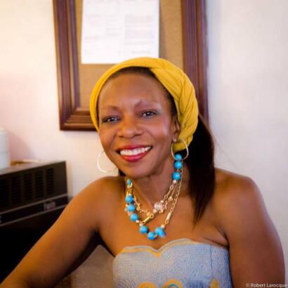 Woman with a yellow headwrap, statement jewelry, and a strapless top smiling in an indoor setting.
