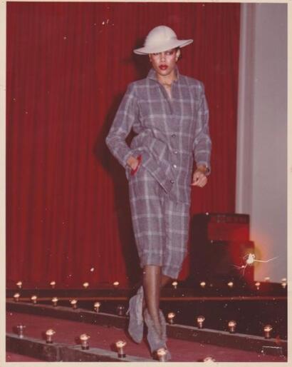 Woman on runway wearing a plaid suit with a wide-brimmed hat, set against red curtains.