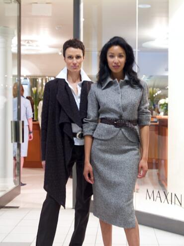 Two women in stylish gray and dark suits pose outside a store.