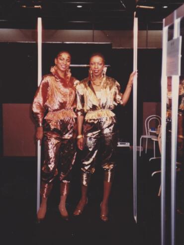 Two women pose in stylish metallic outfits against a dark background.
