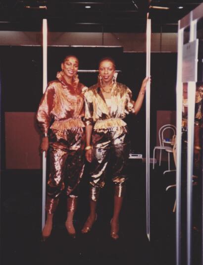 Two women pose in stylish metallic outfits against a dark background.