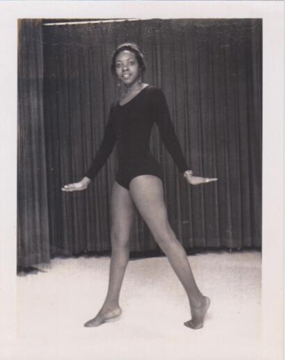 Black and white photo of a young woman in a dance costume, posing confidently against a curtain backdrop.