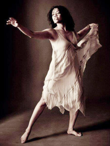 Fashion-forward woman in a flowing beige dress strikes a dynamic pose against a dark background.