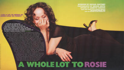 A woman with curly hair and a black outfit, smiling and covering her mouth with her hand, sitting against a yellow background.