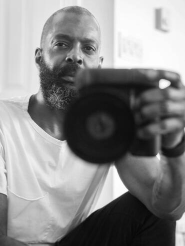 Black-and-white portrait of a bearded man holding a camera, dressed casually, indoors.