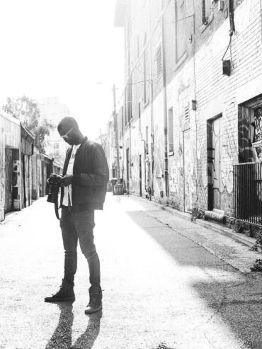 A young man with sunglasses and a camera stands on an urban alleyway, illuminated by bright sunlight.