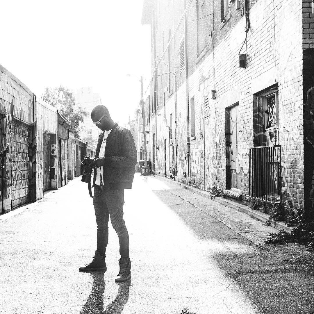 A young man with sunglasses and a camera stands on an urban alleyway, illuminated by bright sunlight.