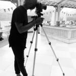 Photographer adjusting camera in bright, open indoor space with kitchen area and decorated ceiling.