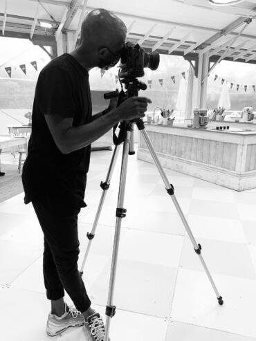 Photographer adjusting camera in bright, open indoor space with kitchen area and decorated ceiling.