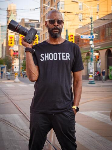 Man wearing a "SHOOTER" T-shirt, sunglasses, and holding a professional camera on a city street.