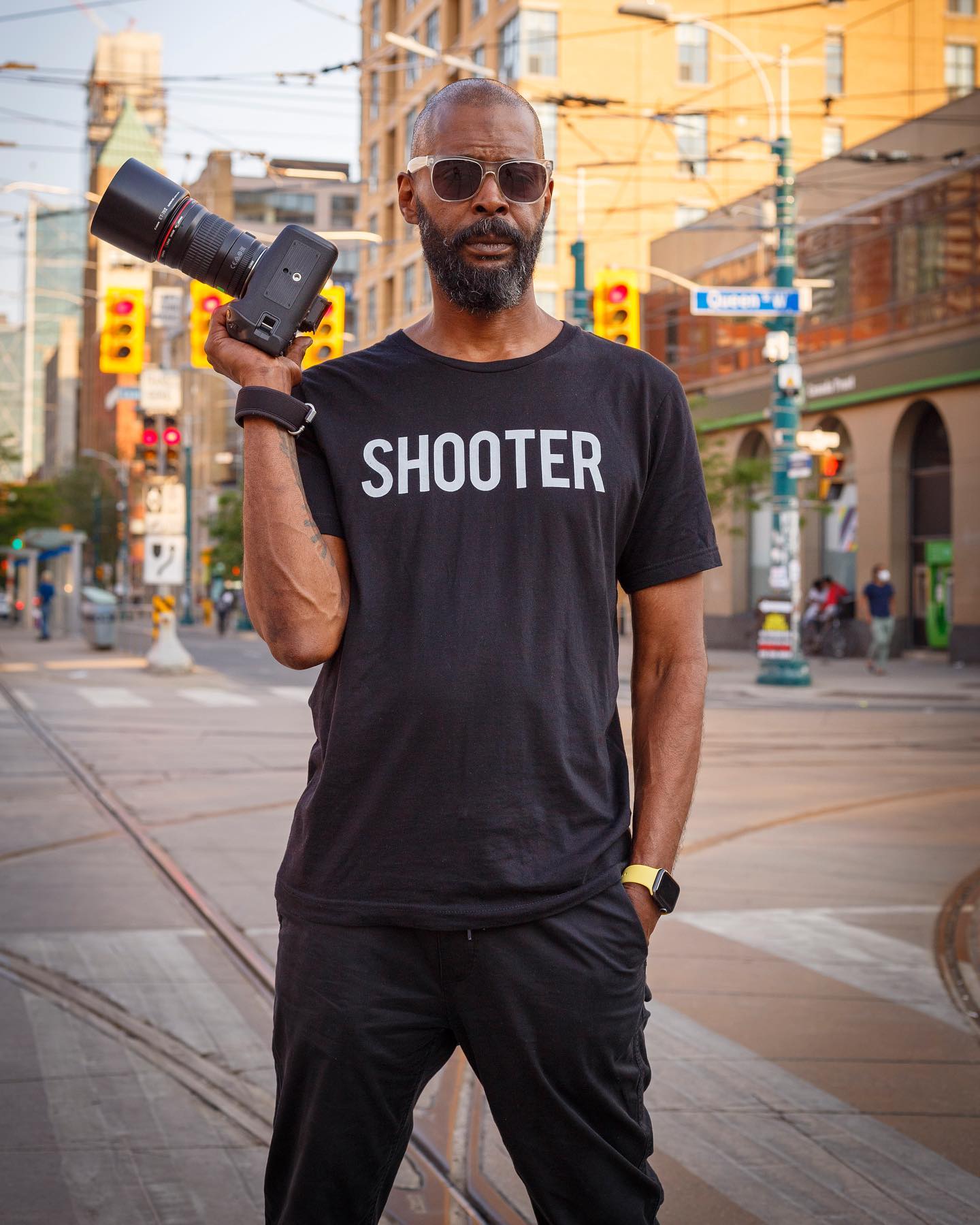 Man wearing a "SHOOTER" T-shirt, sunglasses, and holding a professional camera on a city street.