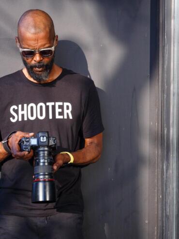 Man with a beard and sunglasses holding a camera against a gray wall.