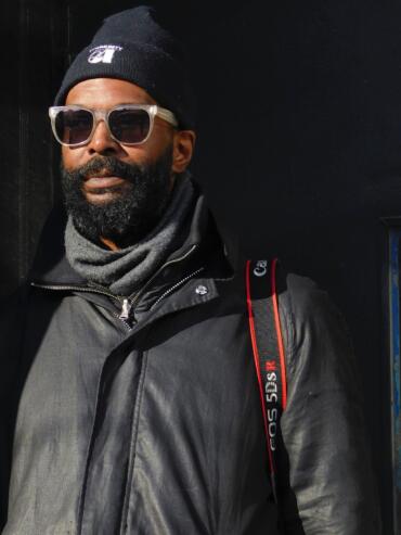 Man with a beard wearing sunglasses, a black beanie, gray scarf, and jacket against a dark background.