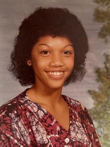 Portrait of a young woman with curly hair wearing a patterned top, smiling against a blurred outdoor background.