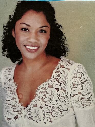 Woman in a white lace top with curly dark hair and a bright smile.