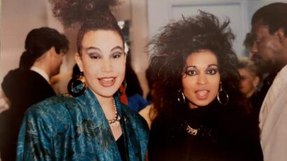 Two women with bold hairstyles and statement jewelry pose at a social event, dressed in vibrant fashion.