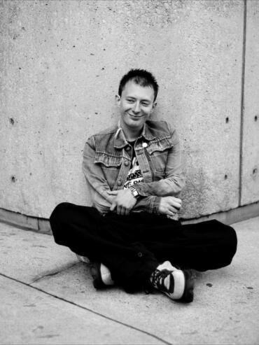 Young man sitting cross-legged against a concrete wall, casual style in denim jacket and sneakers.