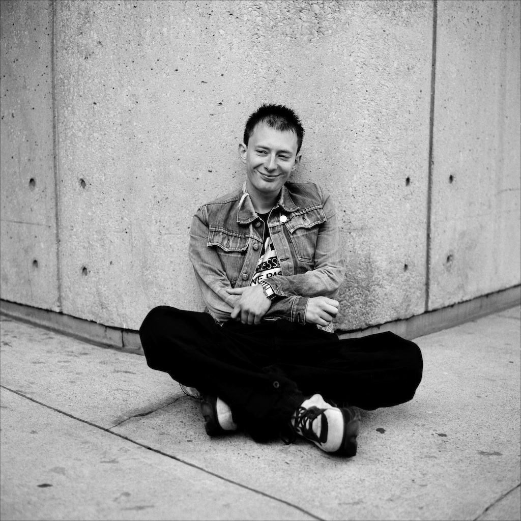 Young man sitting cross-legged against a concrete wall, casual style in denim jacket and sneakers.