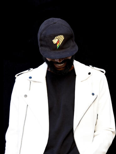 Man wearing a black cap with a lion and rainbow emblem, white jacket, and black shirt against a dark background.