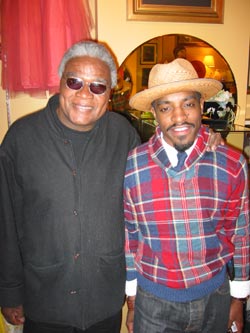 Two men smiling, one in sunglasses and a black jacket, the other in a plaid sweater and straw hat.