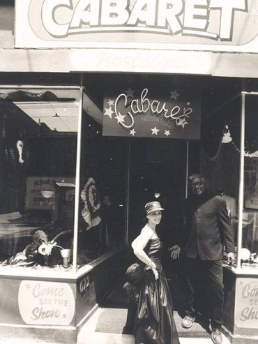 Vintage black-and-white photo of a couple standing outside a cabaret's neon-lit entrance, inviting curiosity.