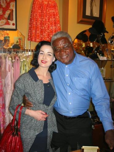 Two stylish individuals, a woman with vintage-inspired makeup and a man in a blue shirt, smile together inside a boutique filled with dresses and accessories.