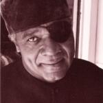 Portrait of an older man with a smiling expression, wearing a hat, an eye patch, and a dark jacket.