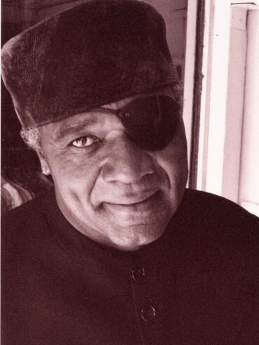 Portrait of an older man with a smiling expression, wearing a hat, an eye patch, and a dark jacket.