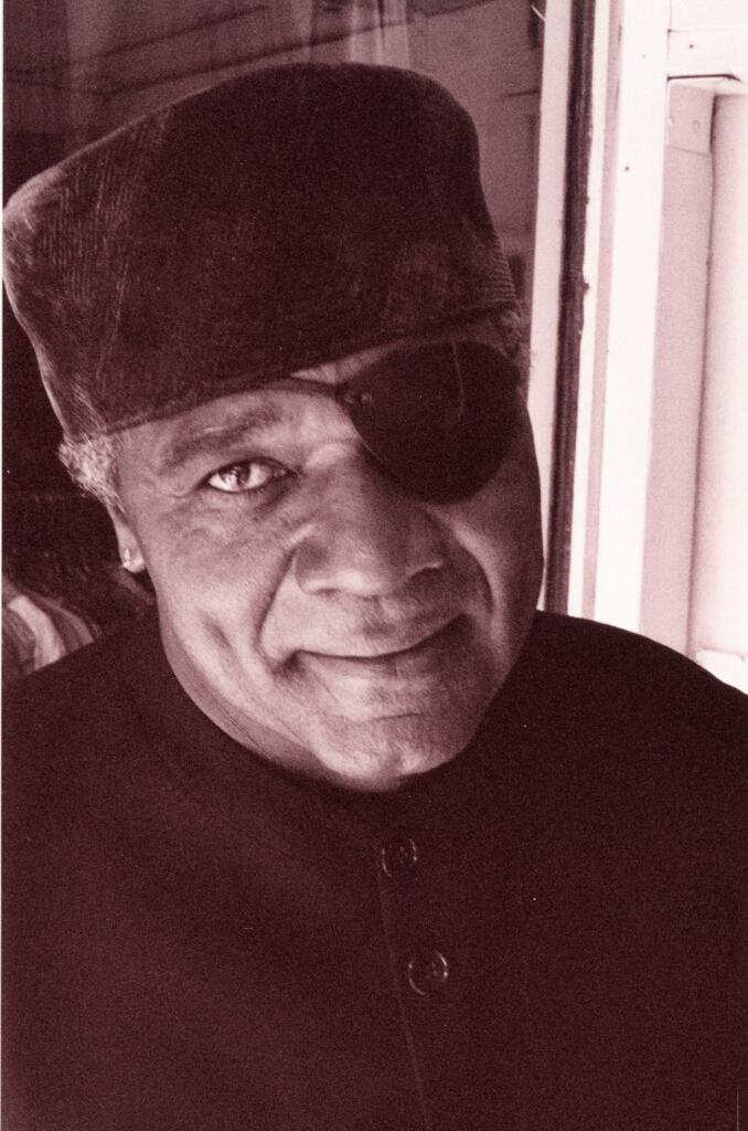Portrait of an older man with a smiling expression, wearing a hat, an eye patch, and a dark jacket.